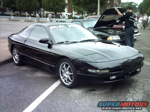Black coupe with chrome wheels parked on asphalt. Another car's hood is open with a person looking inside