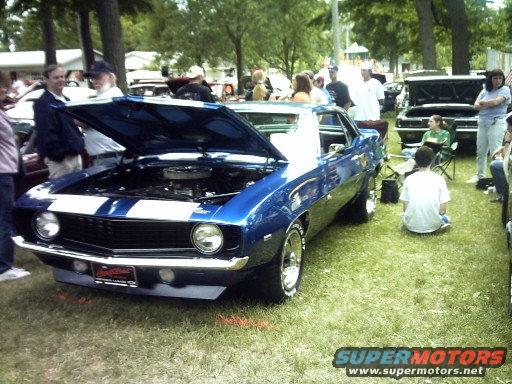 Blue classic car with white racing stripes, hood open, parked on grass at an outdoor event