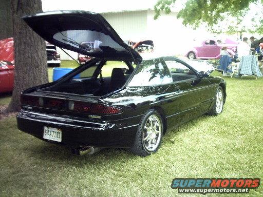 Black coupe with trunk open, parked on grass at an outdoor event. Other cars and people are visible in the background