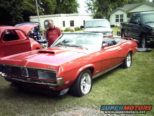 Red convertible classic car with a hood scoop, parked on grass. A man stands nearby. Other vehicles and houses are in the background