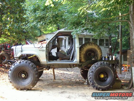 Modified pickup truck with large tires, doors removed, and spare tire mounted in the bed. Another vehicle is partially visible to the left
