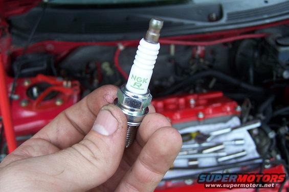 Spark plug with NGK R logo held in hand, engine bay in background