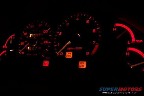 Car dashboard gauges with illuminated red needles and orange warning lights for brake, check engine, and fuel cutout