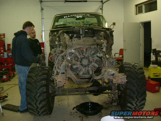 Truck in garage, hood off, engine exposed, big tires, two guys looking at it
