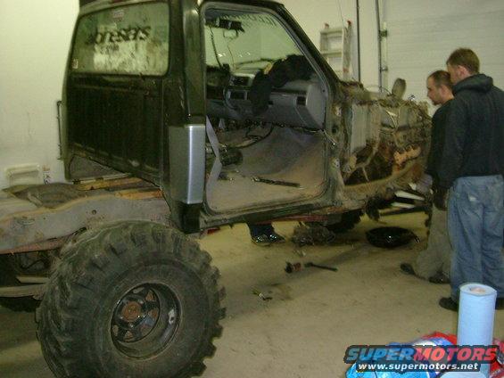 Dark green truck cab and chassis with large tires, missing engine and front clip. Interior visible. Two men stand near the engine bay
