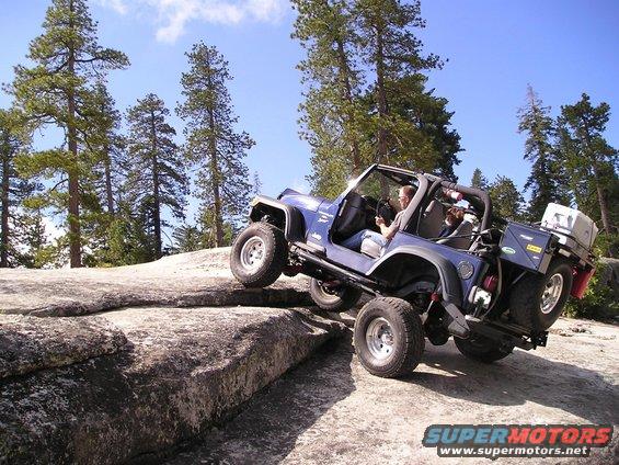 Blue Jeep Wrangler climbing over rocks on a trail, driver visible. Some storage in rear