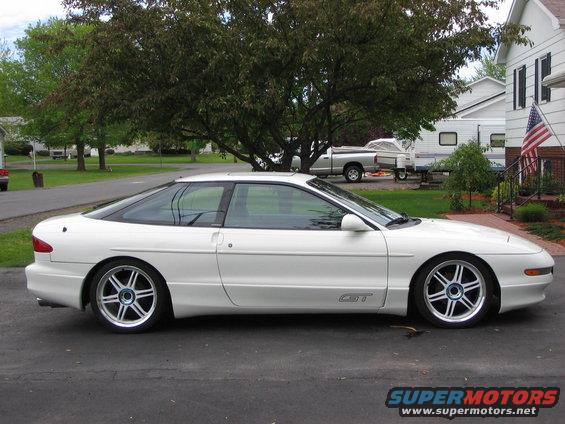 White 1993 Ford Probe GT coupe parked on asphalt, side view. Aftermarket wheels, slight lowering. A pickup truck and trailer are visible in the background