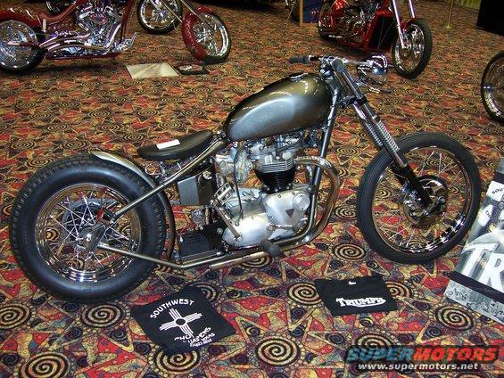 Custom motorcycle with a grey tank and chrome accents, parked on a patterned carpet. Other motorcycles are visible in the background