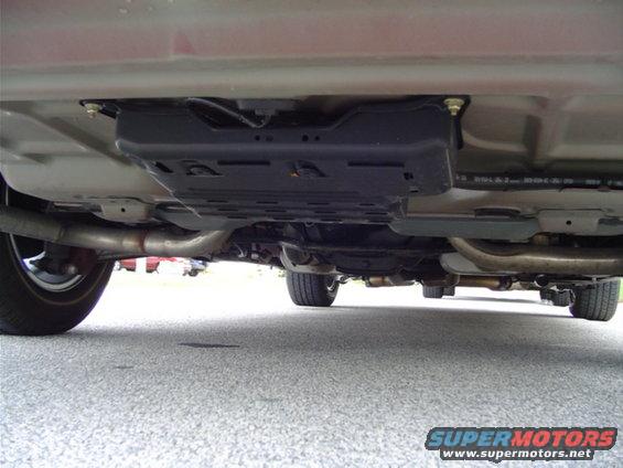 Undercarriage of a red car, showing exhaust pipes and a black plastic shield. The view is from below, looking forward