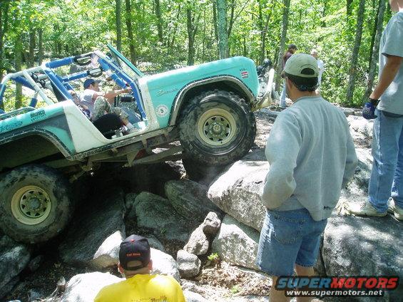 Light blue off-road vehicle with roll cage climbing over large rocks, people watching
