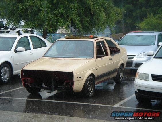 Beige Volkswagen Jetta, missing front bumper and grille, parked in rain. Roll cage visible inside. Other cars nearby