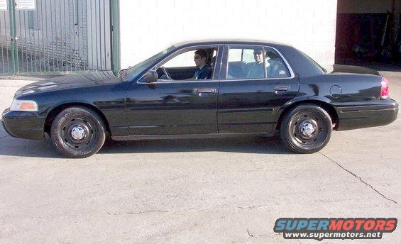 Black 2003 Ford Crown Victoria sedan parked on concrete, side view. Two people inside
