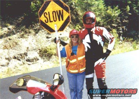 Red motorcycle fairing and mirrors in foreground, two people and a slow sign behind