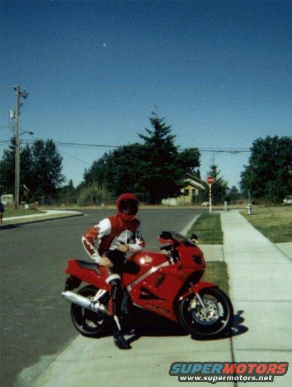 Red motorcycle with rider in red and white gear, parked on sidewalk