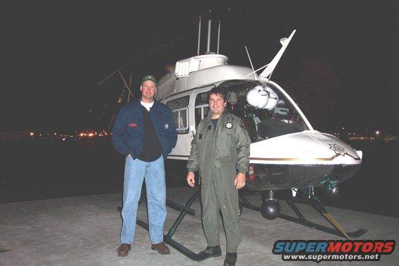 Two men stand next to a white helicopter at night. One wears a jacket and jeans, the other a flight suit
