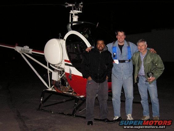 Three men stand in front of a helicopter at night. The helicopter has registration N269SF