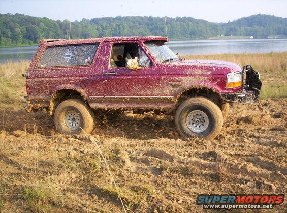 Mud-covered red 1995 Ford Bronco driving through mud near a lake