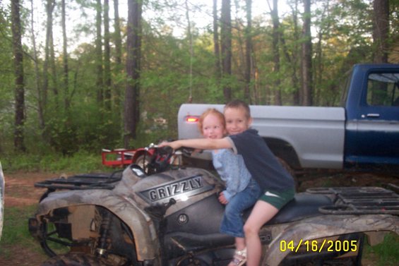 Two kids on a GRIZZLY ATV. A blue and silver pickup truck is behind them