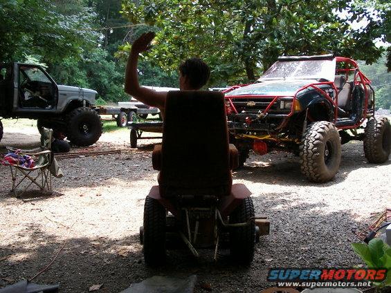 Person on riding mower waves, with a heavily modified off-road vehicle and a lifted truck in the background