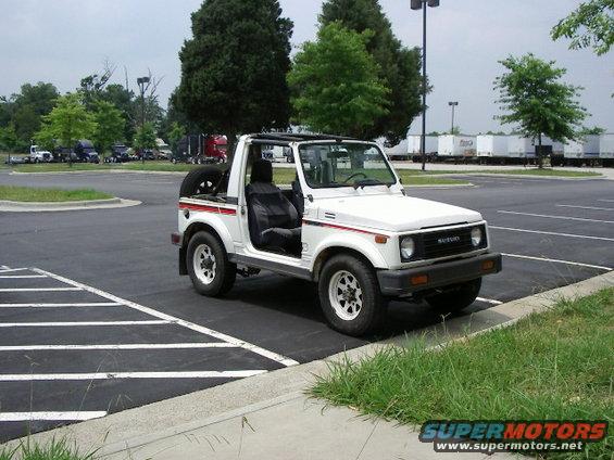 White Suzuki Samurai parked in a lot. It has a spare tire on the back and the driver's door is open