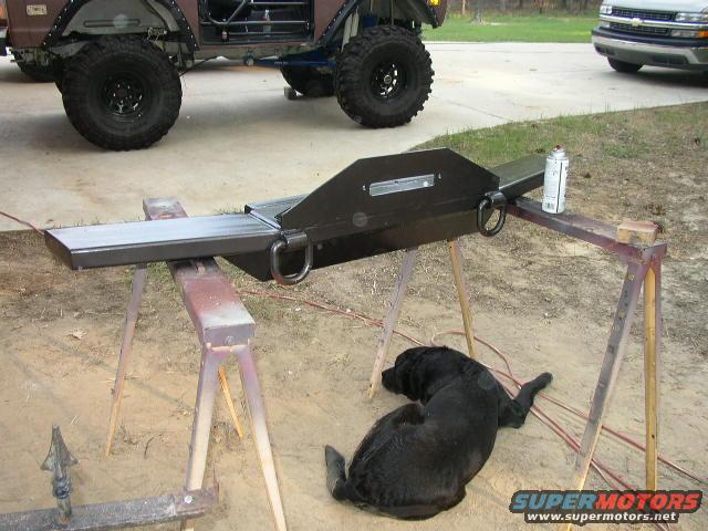 Custom bumper on sawhorses, black dog resting nearby, another vehicle in background