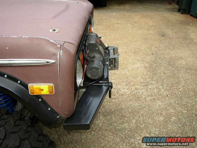 Front corner of a brown Ford Bronco with a winch mounted on the bumper. Shows the headlight, turn signal, and fender flare