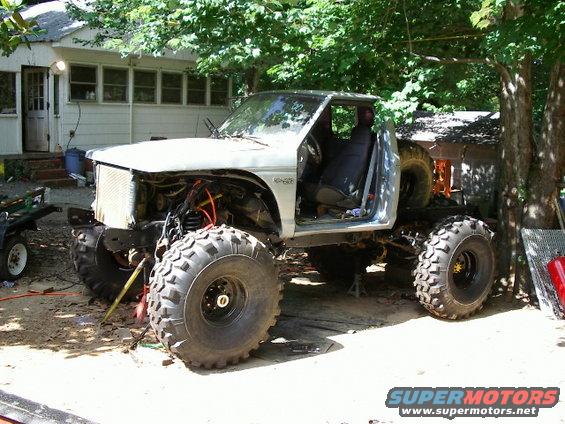 Silver pickup truck with large off-road tires, missing front grille and hood, parked outdoors