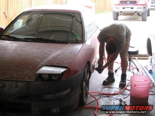 Red 1994 Ford Probe with pop-up headlights and primer, being sanded by a person in a garage