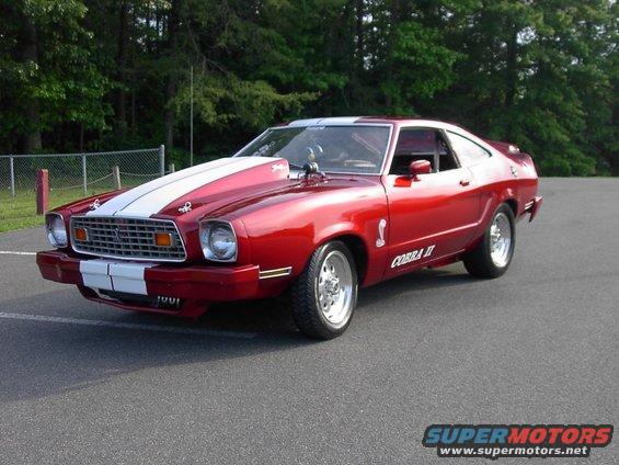 Red 1976 Ford Mustang II Cobra II with white racing stripes on hood and front bumper, chrome wheels, and a hood scoop