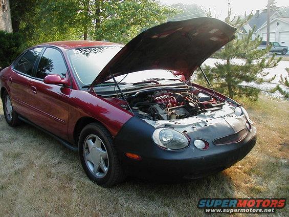 Red sedan with hood open, showing engine and front bumper cover. Grass and trees in background