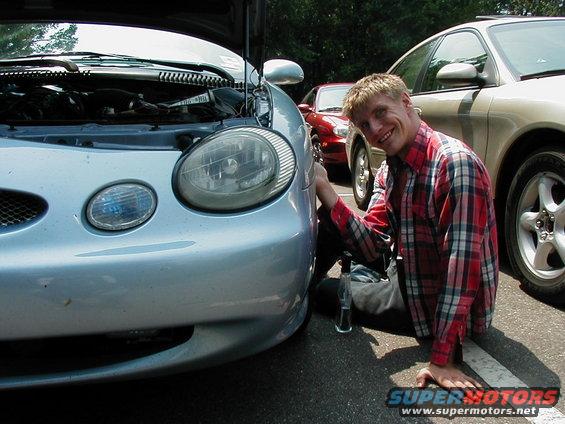 Man in red plaid shirt kneeling by front of light blue 1997 Ford Taurus with hood open; another car visible behind