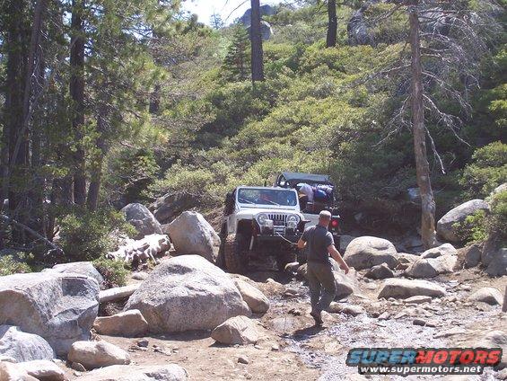 White Jeep Wrangler with winch on a rocky, wooded trail, with another Jeep behind it