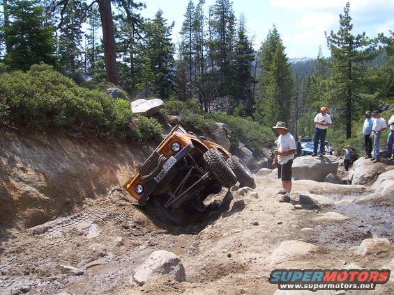 Orange off-road vehicle tilted on its side on a rocky trail with people watching