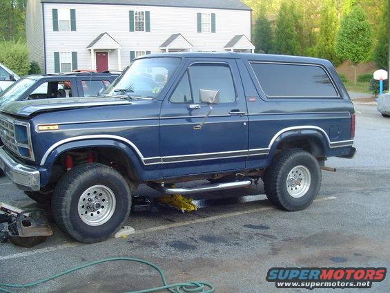 Blue SUV on jack stands with front end partially disassembled. Lifted suspension and aftermarket wheels are visible