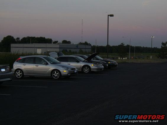silver Ford Focus hatchback, silver sedan with hood up, dark sedan, and part of a gray car in a parking lot at dusk