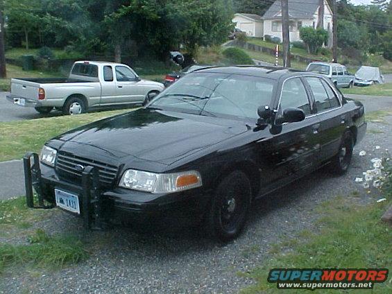 Black police car with push bar and light bar, parked on gravel. License plate visible