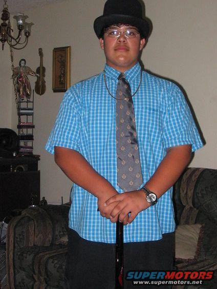 Young person wearing a bowler hat, blue plaid shirt, and tie, holding a cane. Room interior visible