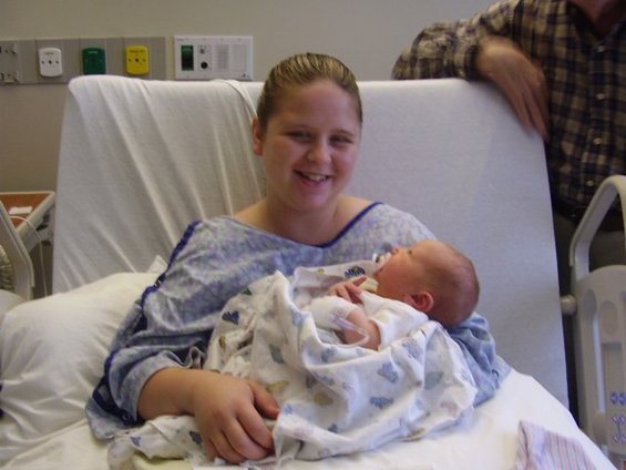 Woman in hospital gown holding a swaddled newborn baby, with a person in a plaid shirt behind her
