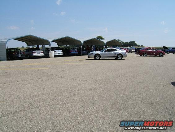 Silver coupe parked on asphalt, side view. Multiple cars under canopies in background