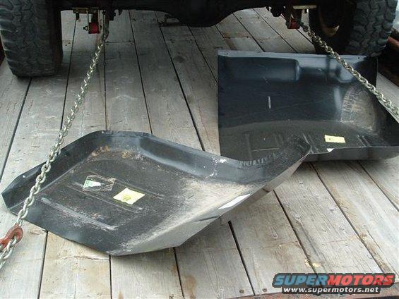 Two black metal floor pans, one curved and one flat, resting on wooden planks. Chains are attached to the underside of a vehicle above