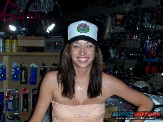 Woman wearing a white trucker hat with a circular logo, smiling in a store with shelves of merchandise