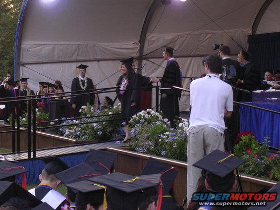 People in graduation gowns on a stage, some receiving leis. Audience in foreground wearing graduation caps