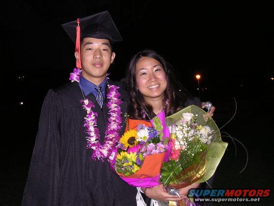 Two people, one in graduation cap and gown with leis, the other holding bouquets of flowers