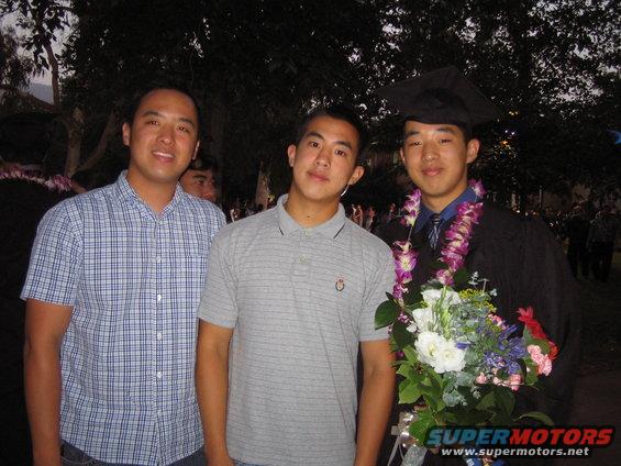 Three men posing for a photo at a graduation ceremony. One is wearing a cap and gown with leis