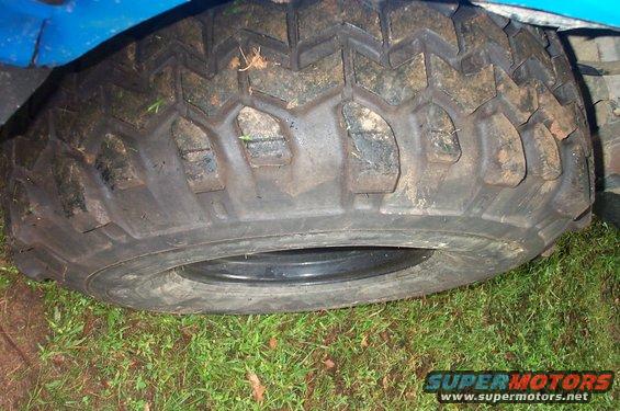 Close-up of a large, knobby off-road tire on a vehicle. The tire is dirty and has a deep tread pattern. Part of the blue body of the vehicle is visible above the tire