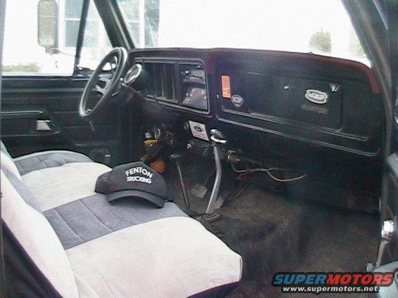 Interior of a truck cab, showing the dashboard, steering wheel, and seats. A hat with 'Fenton Trucking' is on the passenger seat