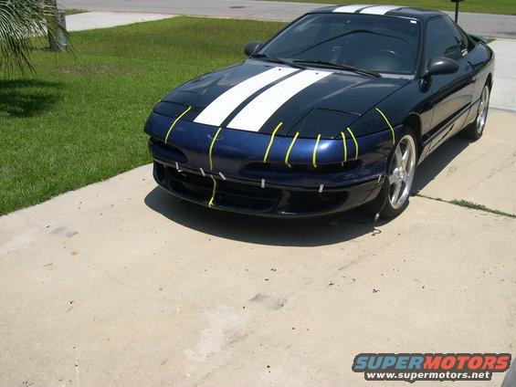 Blue 1997 Ford Probe with white racing stripes and yellow tape on the front bumper, parked on a driveway