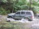 2000 Jeep Cherokee - A Jeep Cherokee navigates a stream in a forest, kicking up water as it goes.