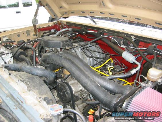 Engine bay with V8 engine, red inner fender, and a large black air intake. A white truck is partially visible in the background