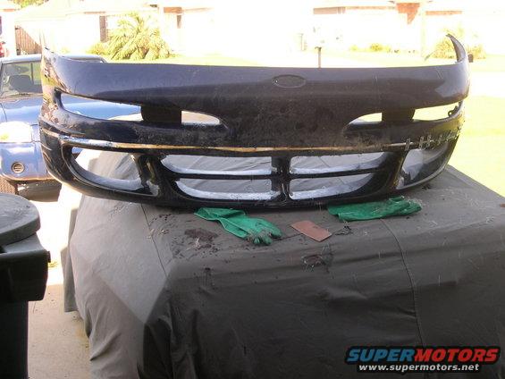 Dark colored car bumper with grille and headlight openings, resting on a covered surface. A blue vehicle is visible in the background